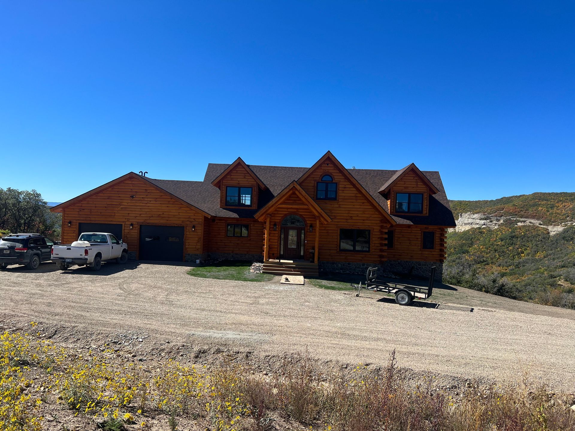 Wooden mountain lodge with garage, parked vehicles, and gravel driveway under a clear blue sky