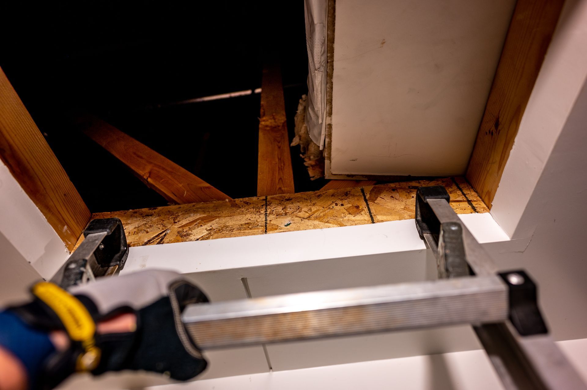 Handyman holding a metal framing square over an exposed ceiling opening with wooden beams and insulation.