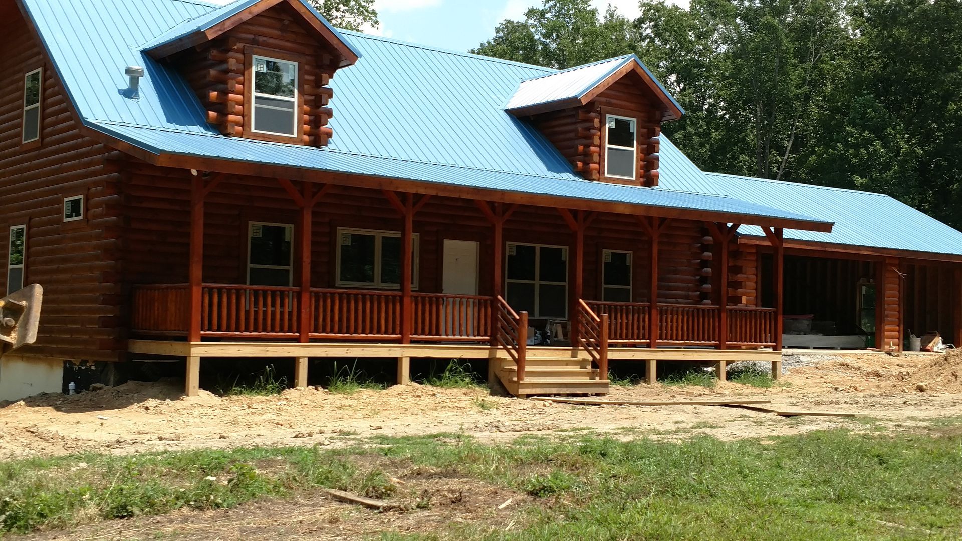 Log cabin with blue metal roof, wraparound porch, and dormer windows on a sandy lot