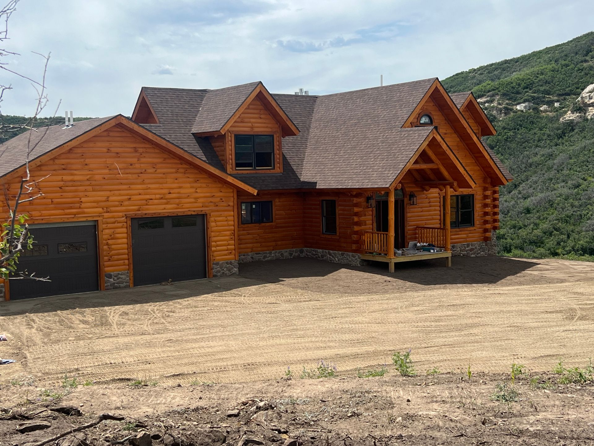Wooden mountain lodge with garage, set on a dirt lot under a cloudy sky