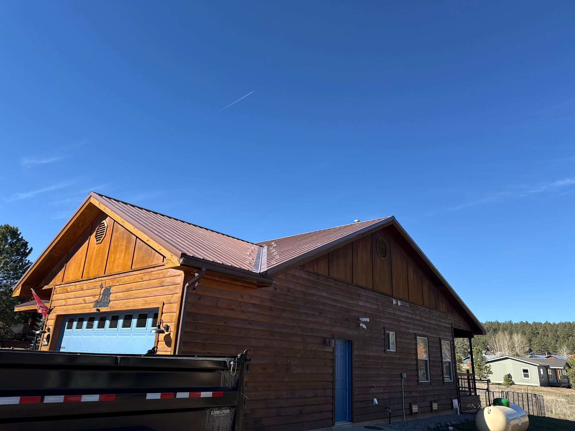 Wooden cabin with brown roof under a clear blue sky, seen beside a black trailer in a rural setting