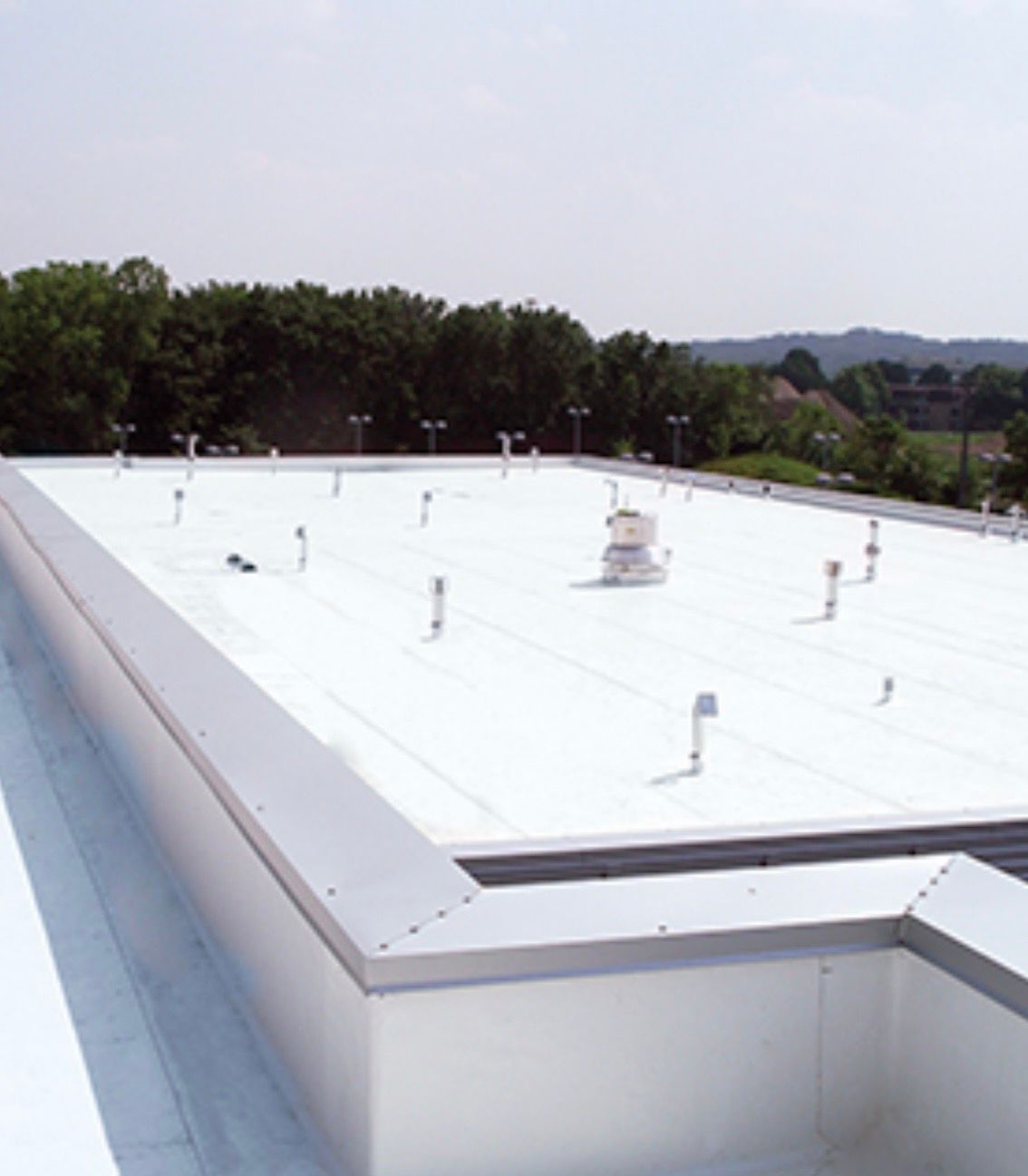 White flat rooftop with vents and a surrounding parapet, viewed against trees and distant hills.
