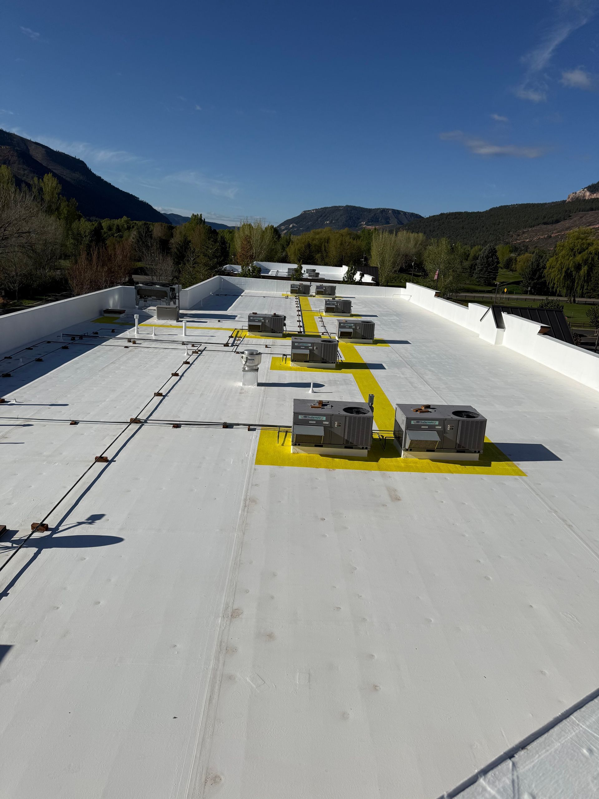 Flat white rooftop with HVAC units and yellow safety markings under a clear blue sky, surrounded by trees and hills