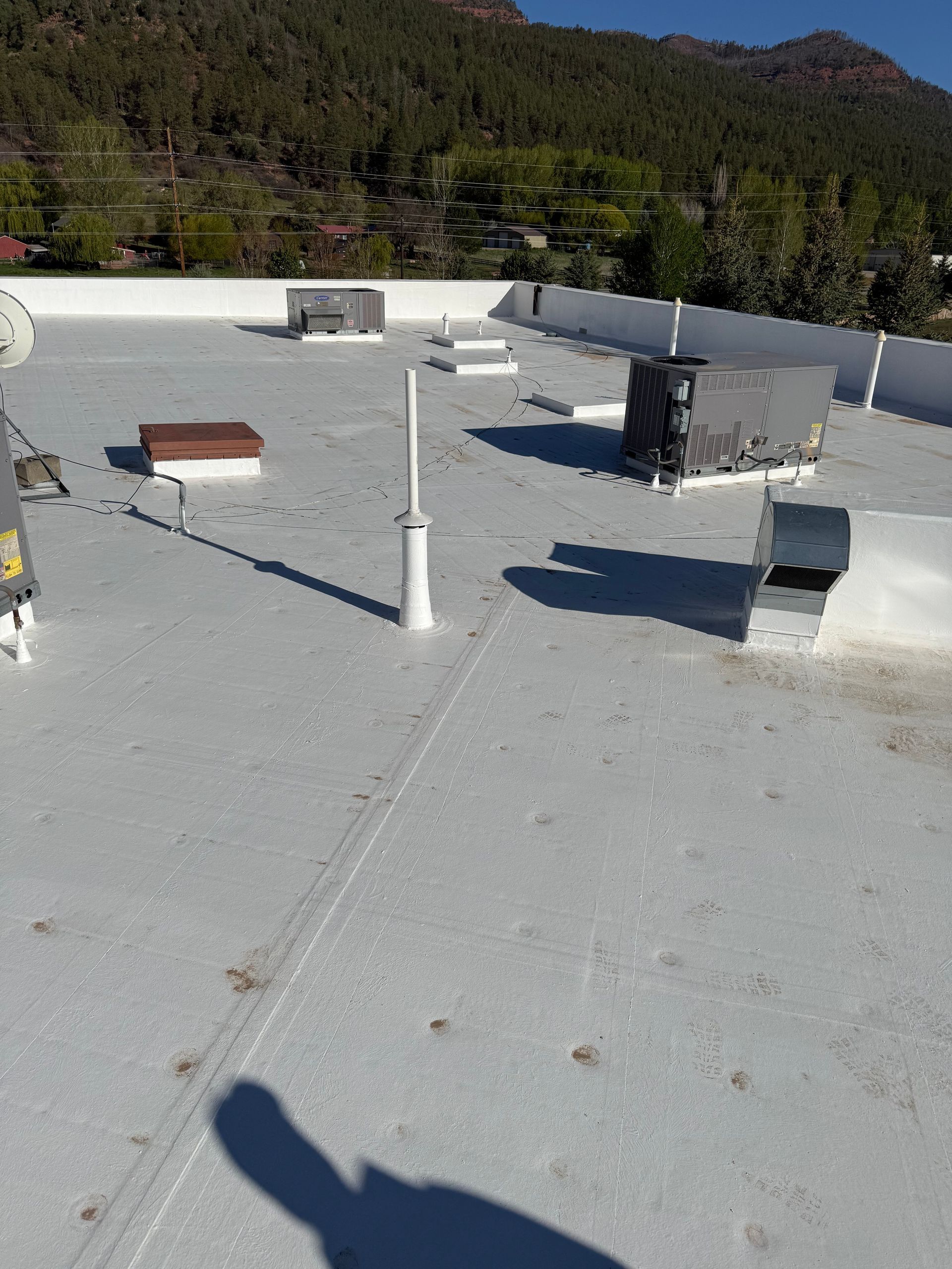 Rooftop with white gravel, vents, pipes, and shadows overlooking a wooded hillside