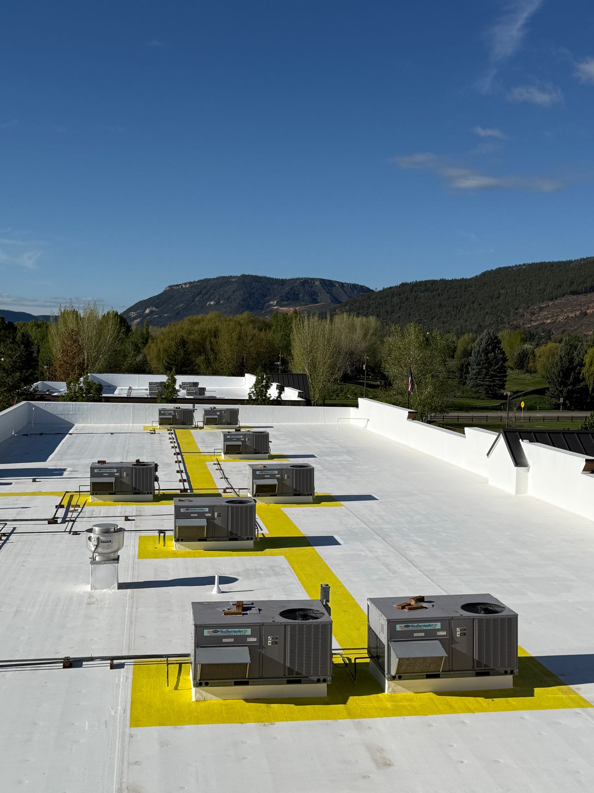 Rooftop HVAC units on a white roof with yellow markings, trees and hills in the background under a blue sky