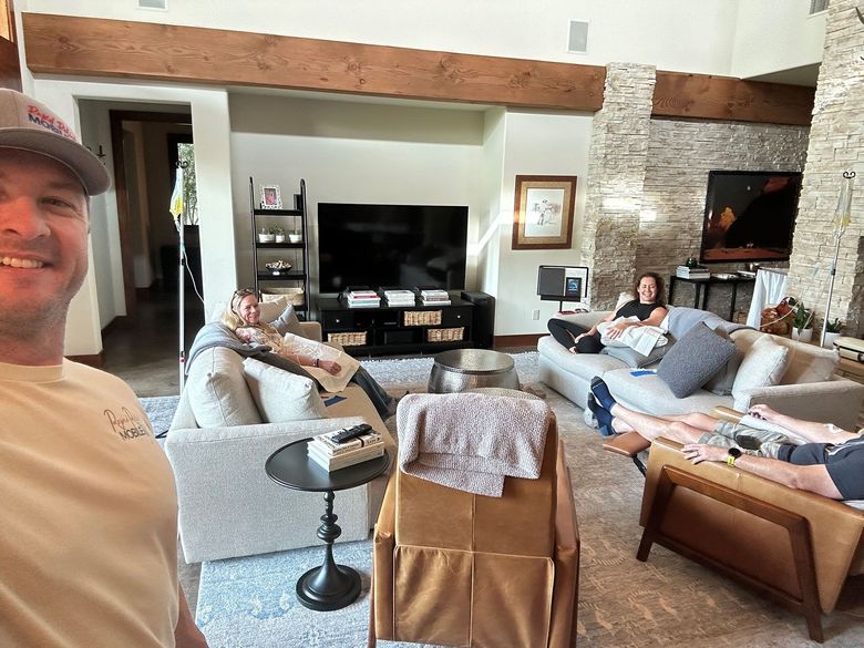 Living room with several people lounging on sofas near a TV and fireplace, viewed in a wide selfie shot