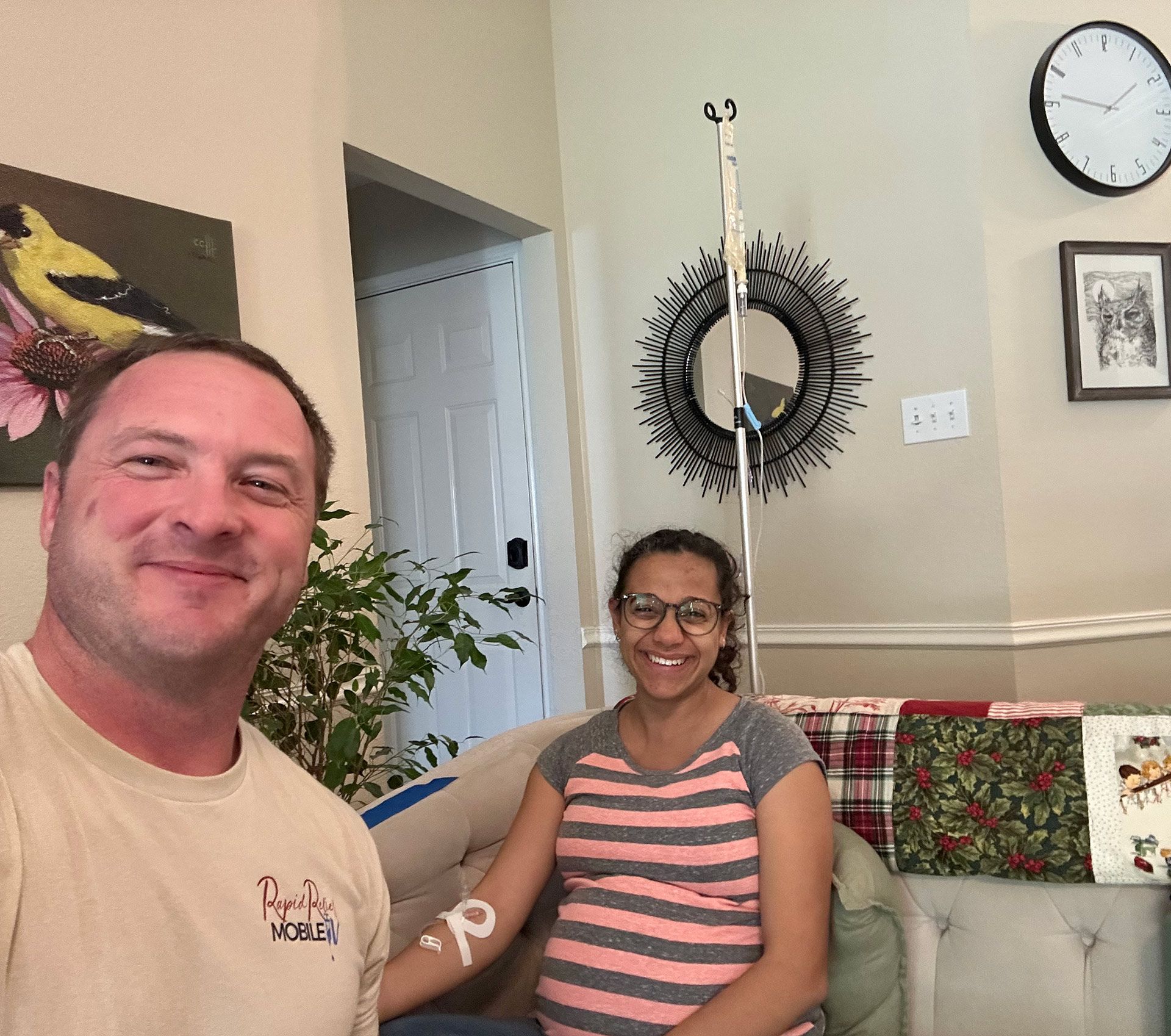 Two people smiling in a living room, one taking a selfie, with wall decor and a clock in the background