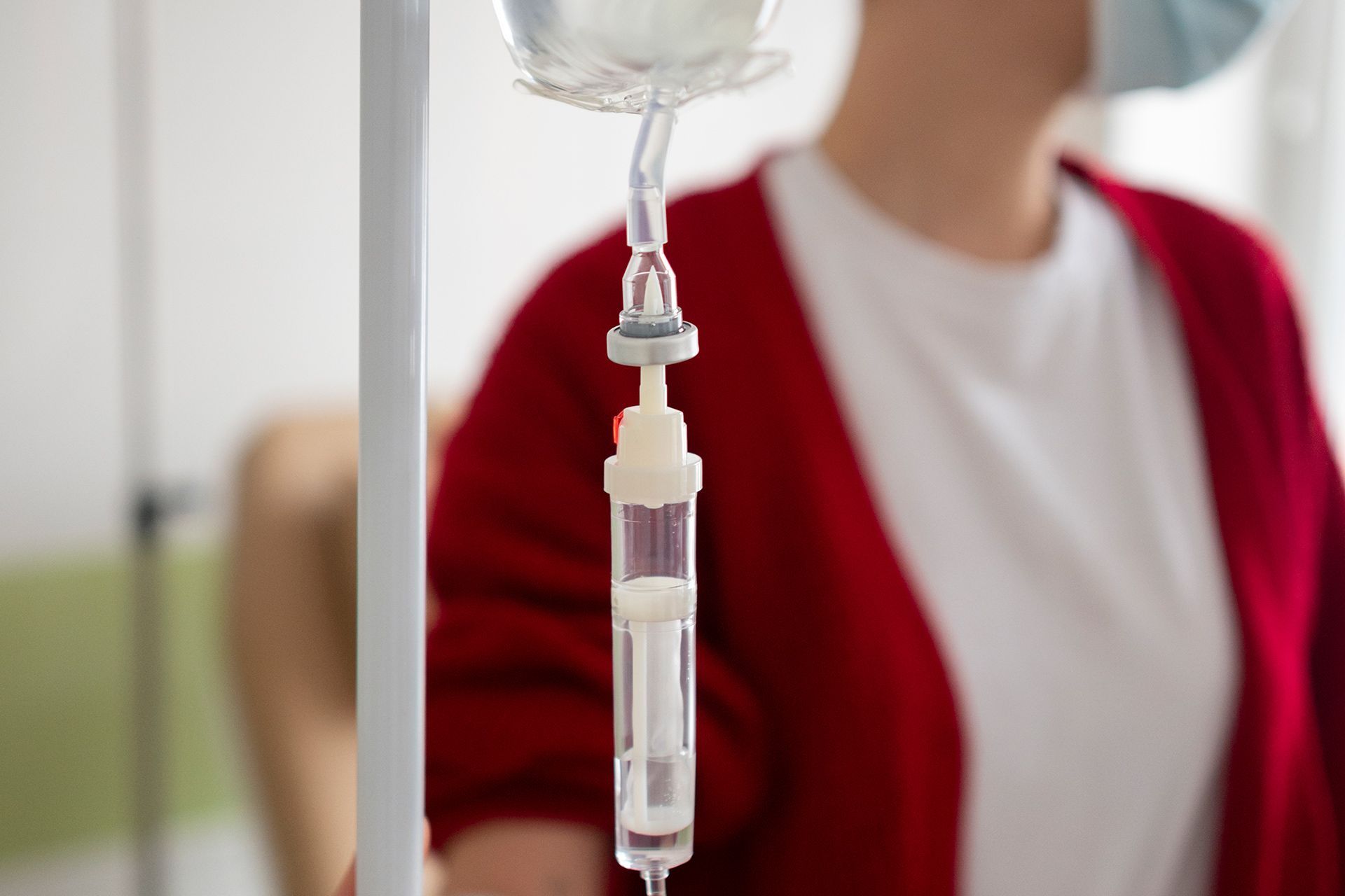 IV drip bag and tubing in foreground, with a blurred person in a red cardigan behind it