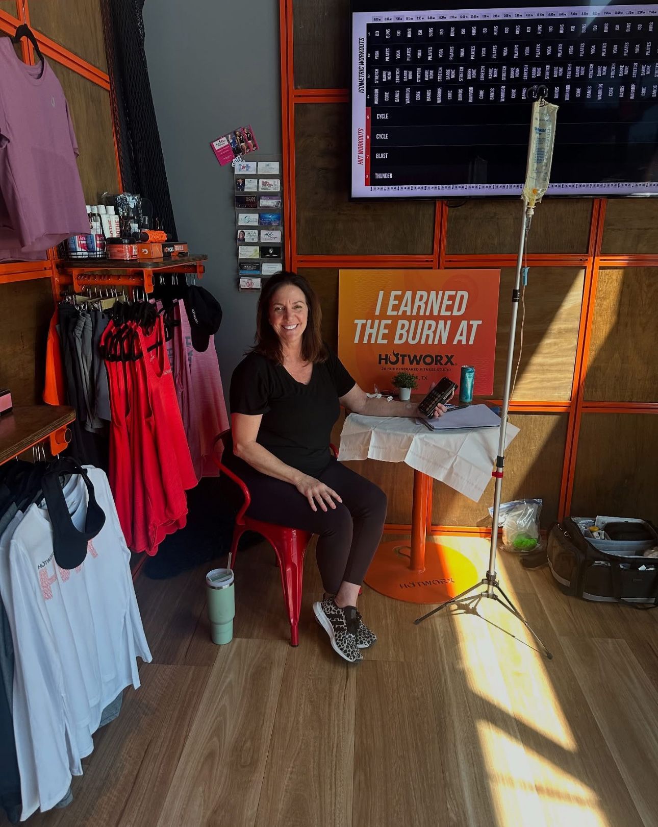 Woman seated in a gym beside a motivational sign, with workout apparel displays and a TV screen.