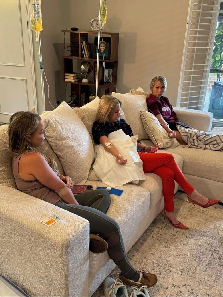 Three people seated on a white couch in a bright living room, one holding a tissue, with pillows and shelves nearby