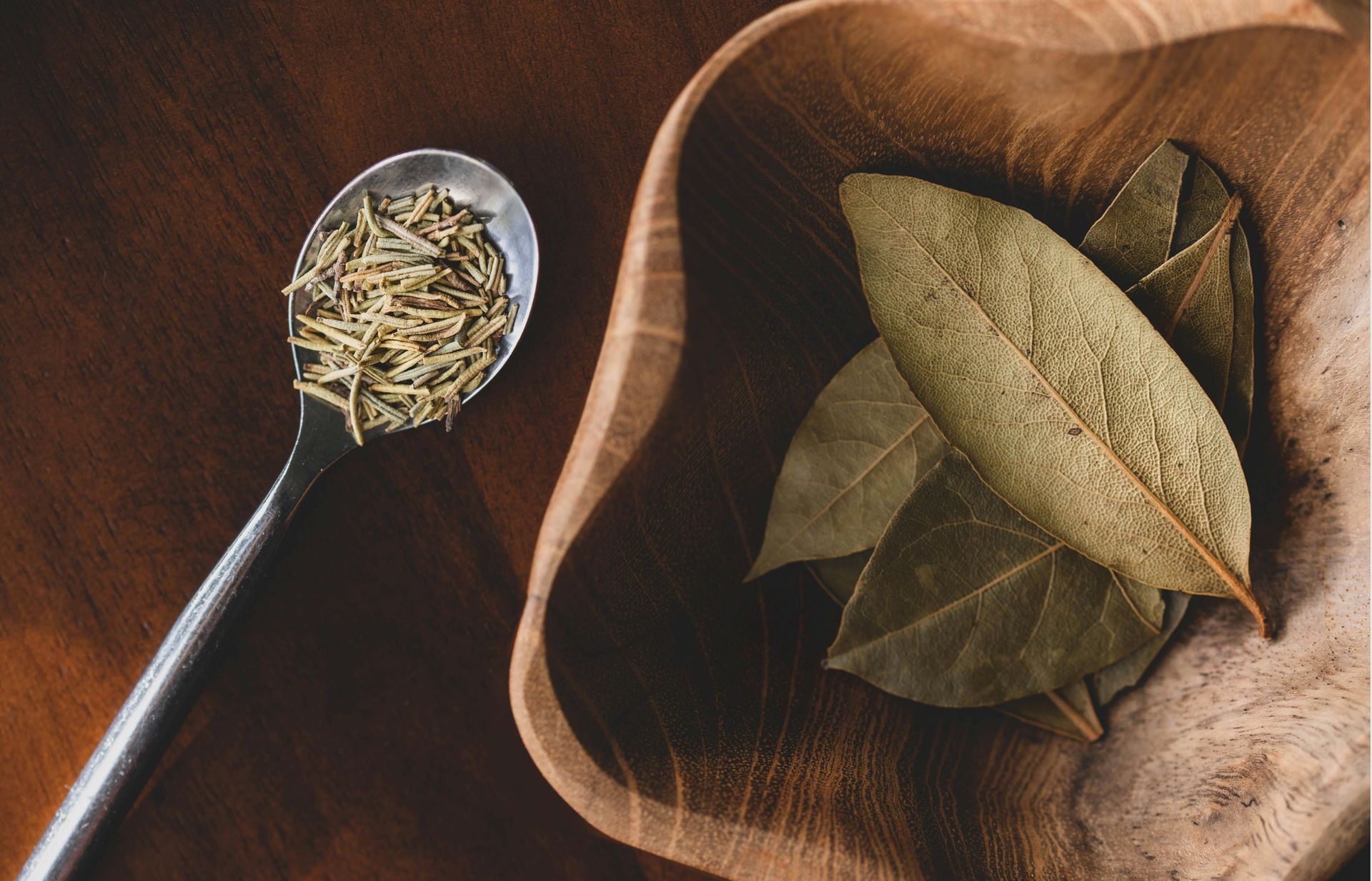 A bowl of bay leaves and a spoon of laurum on a wooden table.