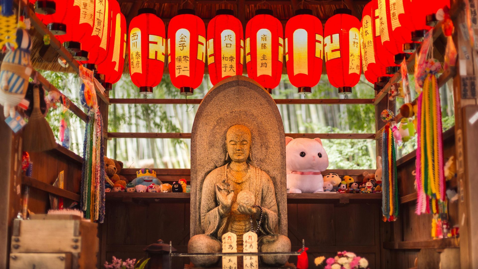 Stone Buddha statue in a shrine, red lanterns overhead, surrounded by trinkets and greenery.