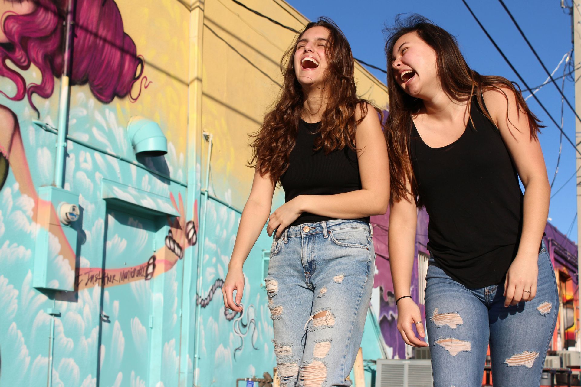 Two young women are laughing in front of a graffiti wall.