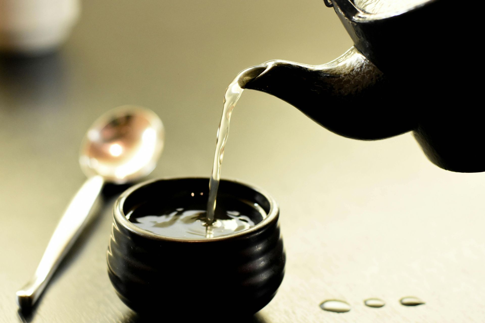 Tea being poured from a dark teapot into a small, dark teacup. A spoon rests nearby.