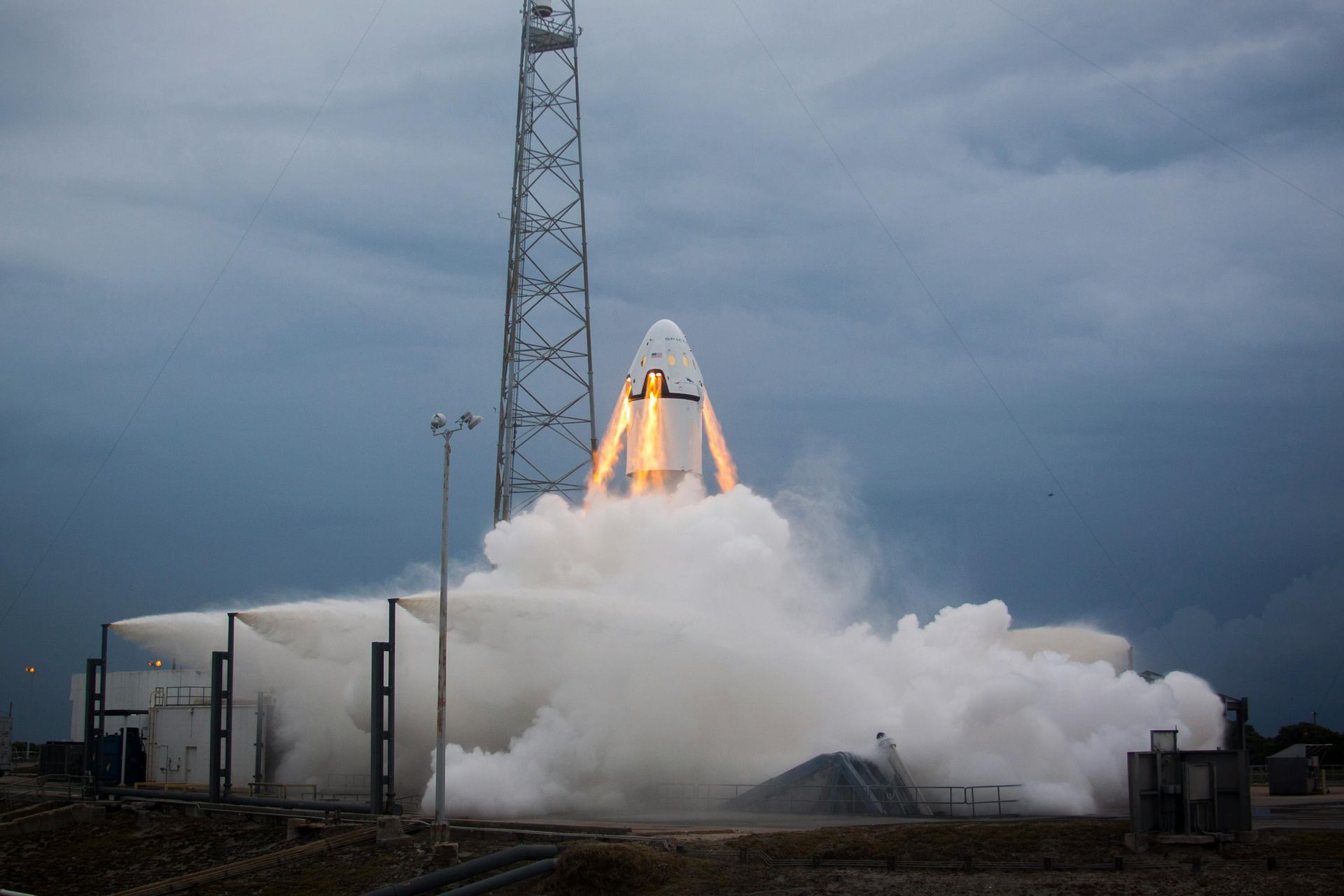 A rocket launches from a pad, flames and smoke billowing, cloudy sky overhead.