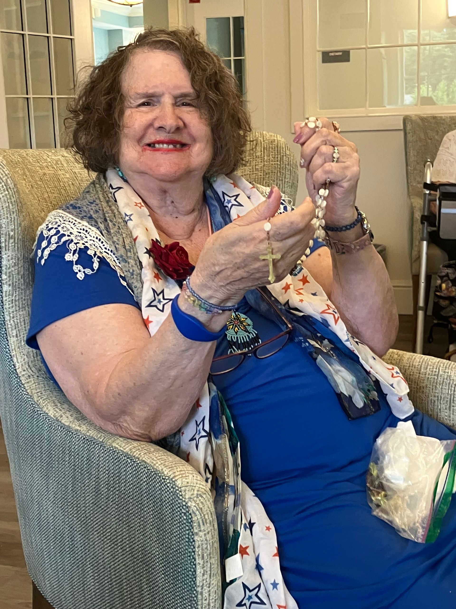 Smiling woman in blue dress and floral shawl holding a beaded necklace while seated indoors