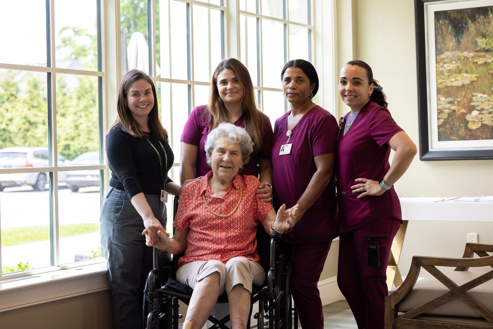 Five women in burgundy scrubs stand beside an elderly woman in a wheelchair by bright windows.