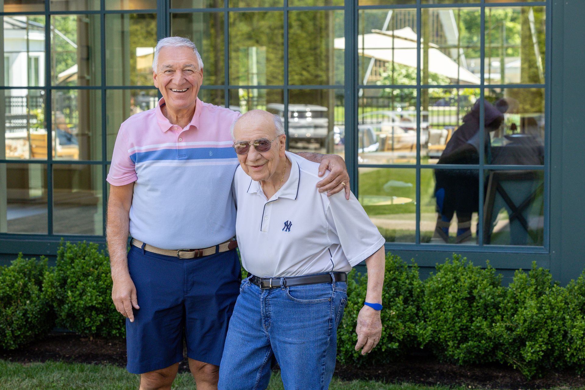 Two smiling people standing outside a building with large windows and green shrubs.