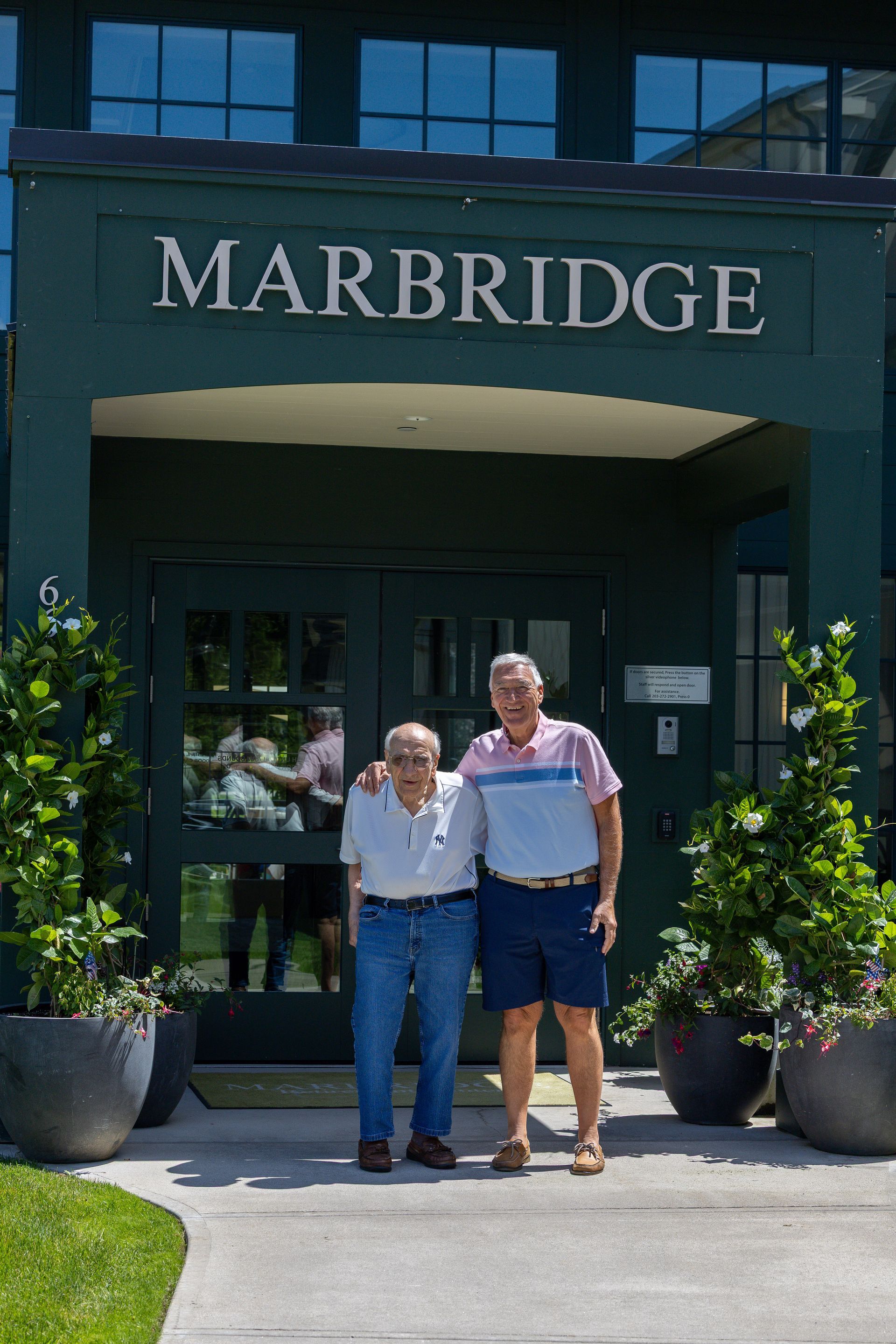 Two people standing outside the MARBRIDGE entrance, with potted plants and a green awning.