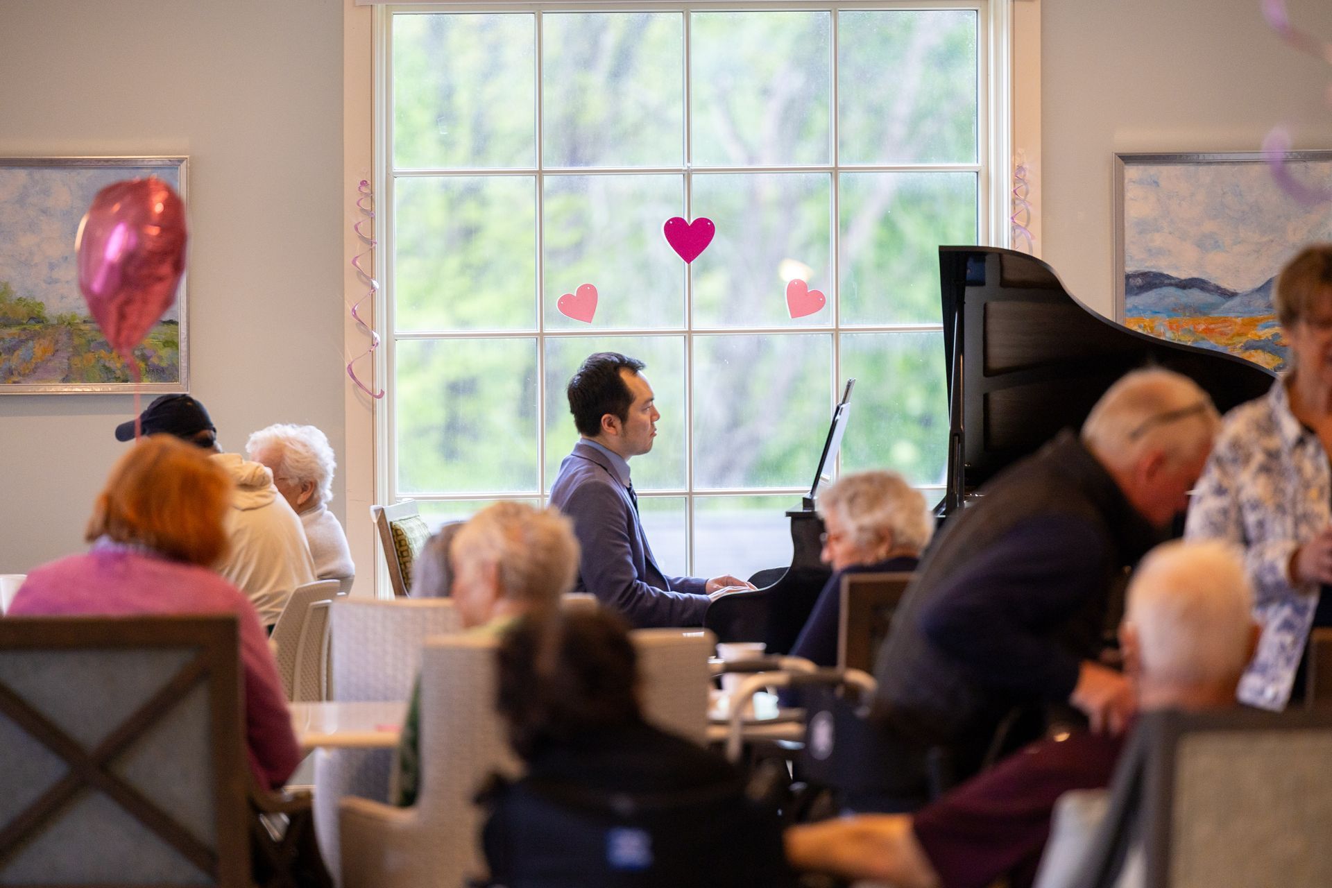 People seated in a room watching a man playing a grand piano by a large window with heart decorations.