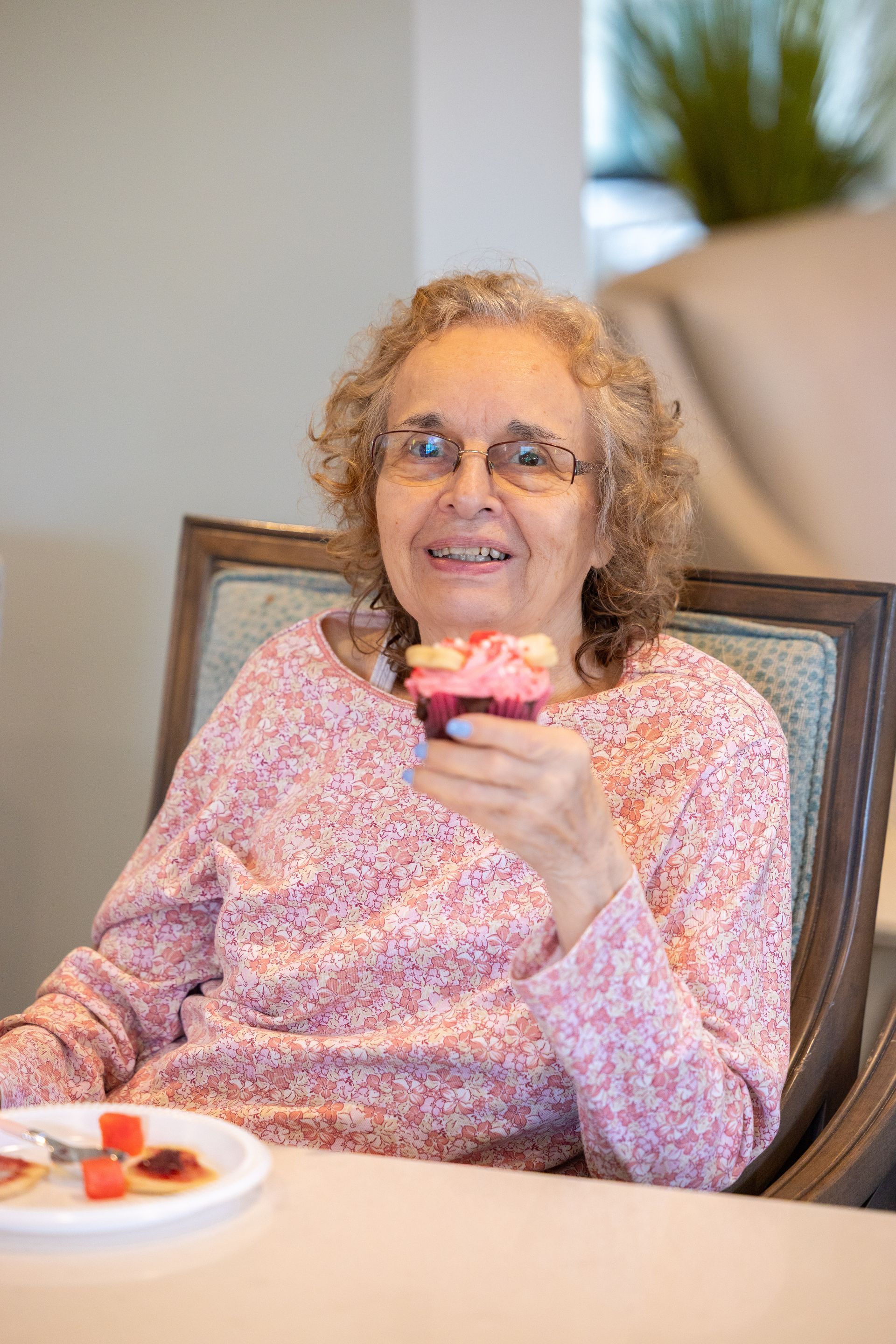 Woman smiling on a couch, holding a cupcake in a bright living room.