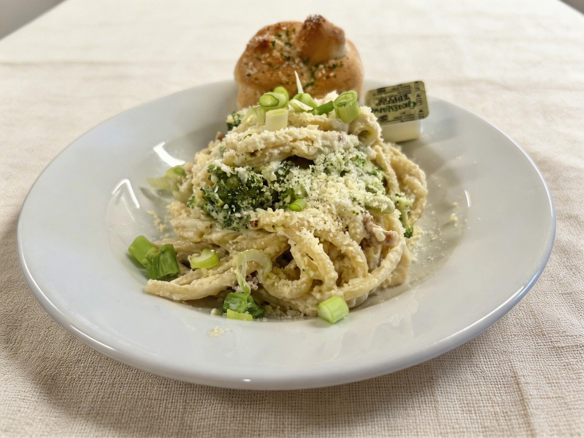 Plate of pasta topped with grated cheese and greens, with bread on a white tablecloth