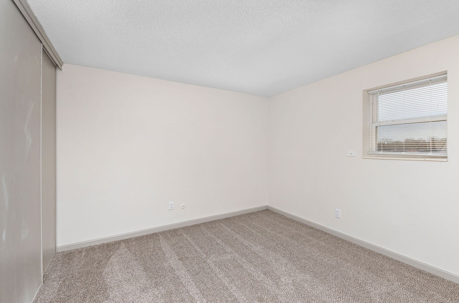 Empty beige bedroom with carpeted floor, white walls, and a small window with blinds.
