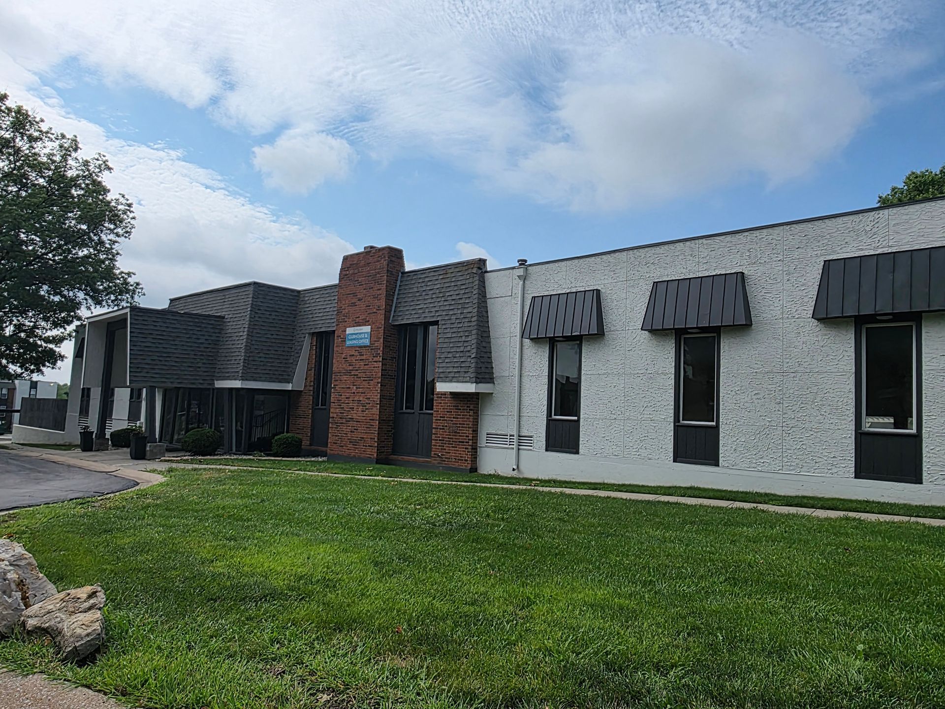 Single-story gray brick building with dark awnings, a red brick entrance, and a grassy lawn under a cloudy sky
