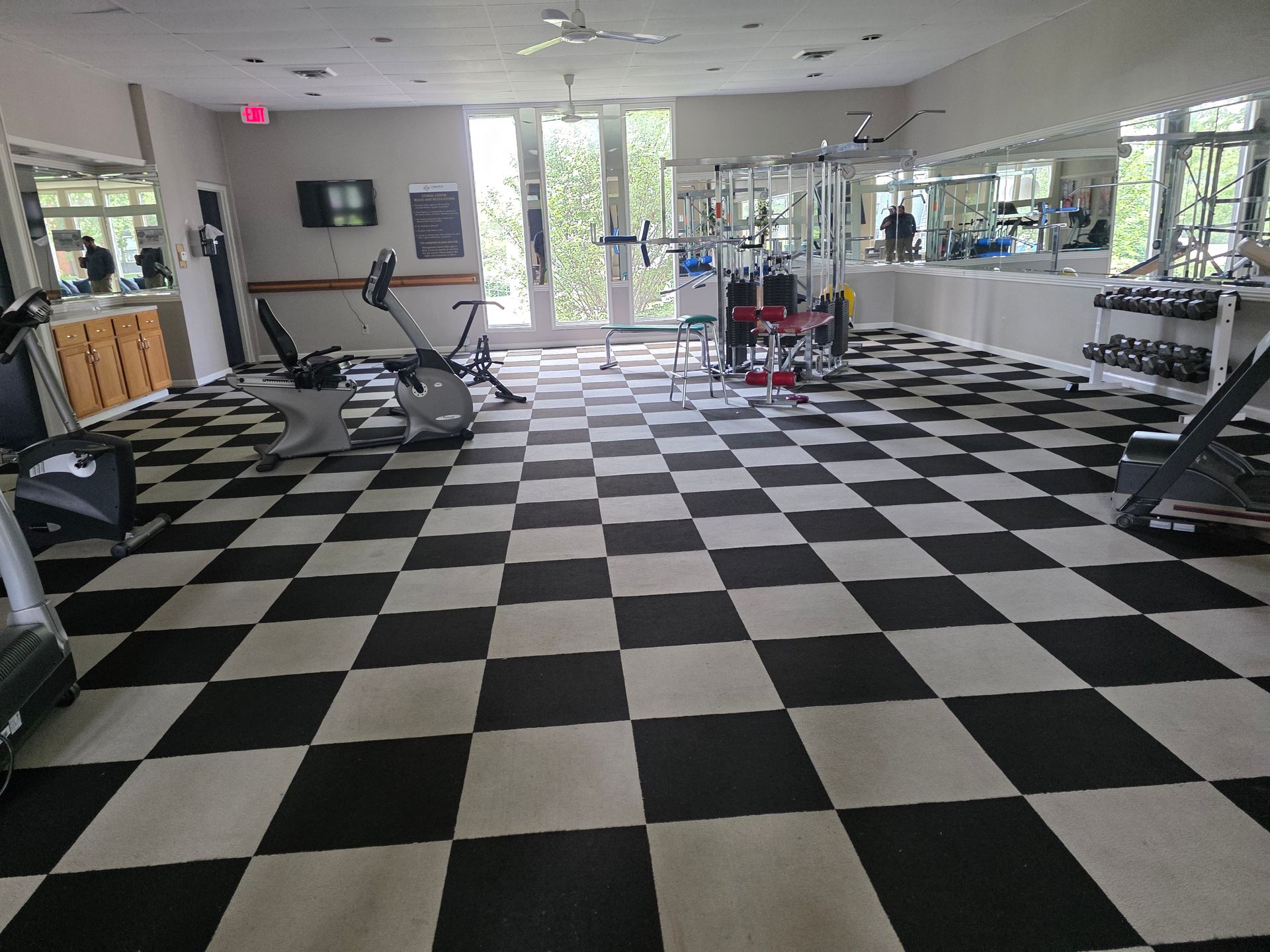 Empty gym with black-and-white checkered floor, treadmills, benches, mirrors, and windows letting in daylight