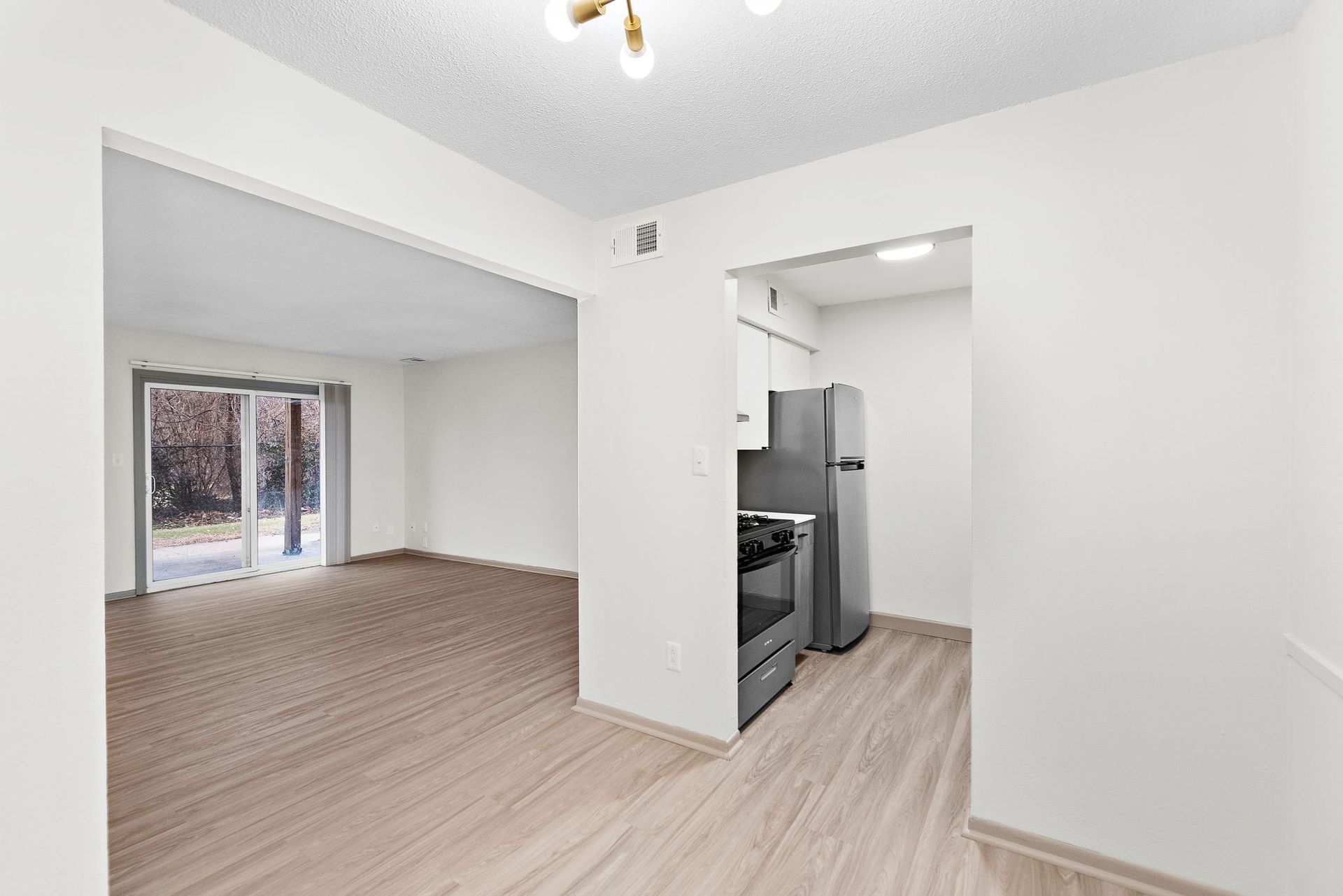 Empty apartment living room and kitchen with white walls, light wood floors, and sliding glass doors to a balcony