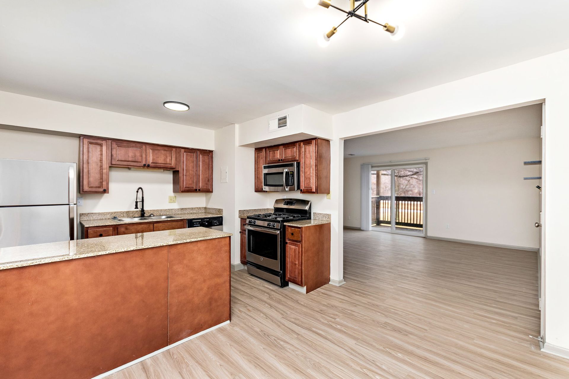 Open-plan kitchen with wood cabinets, stainless fridge, and a view into an empty living area