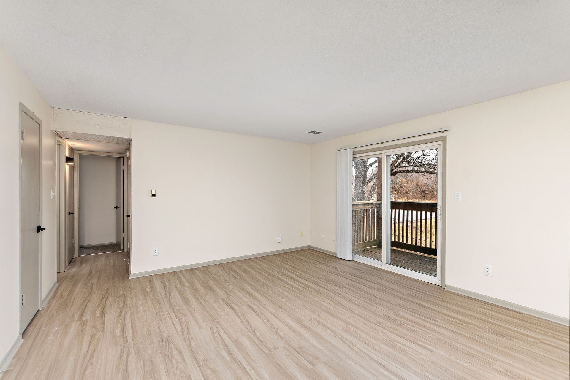 Empty bright living room with light wood floors, white walls, and sliding glass doors to a balcony.