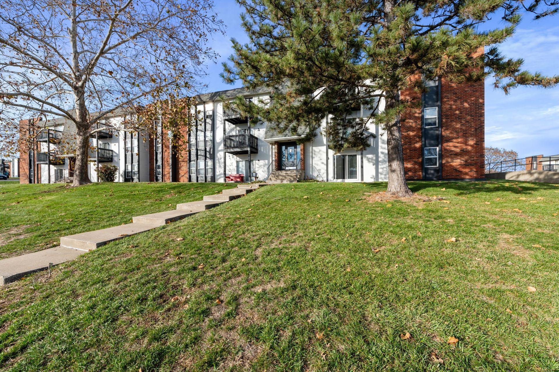 Apartment building with green lawn, concrete walkway, and trees on a sunny day