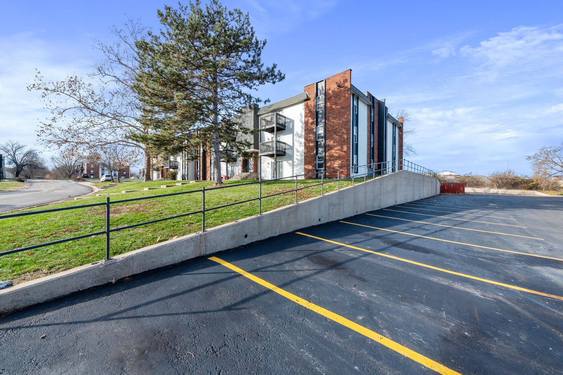 Empty parking lot beside a modern apartment building and grassy slope under a blue sky