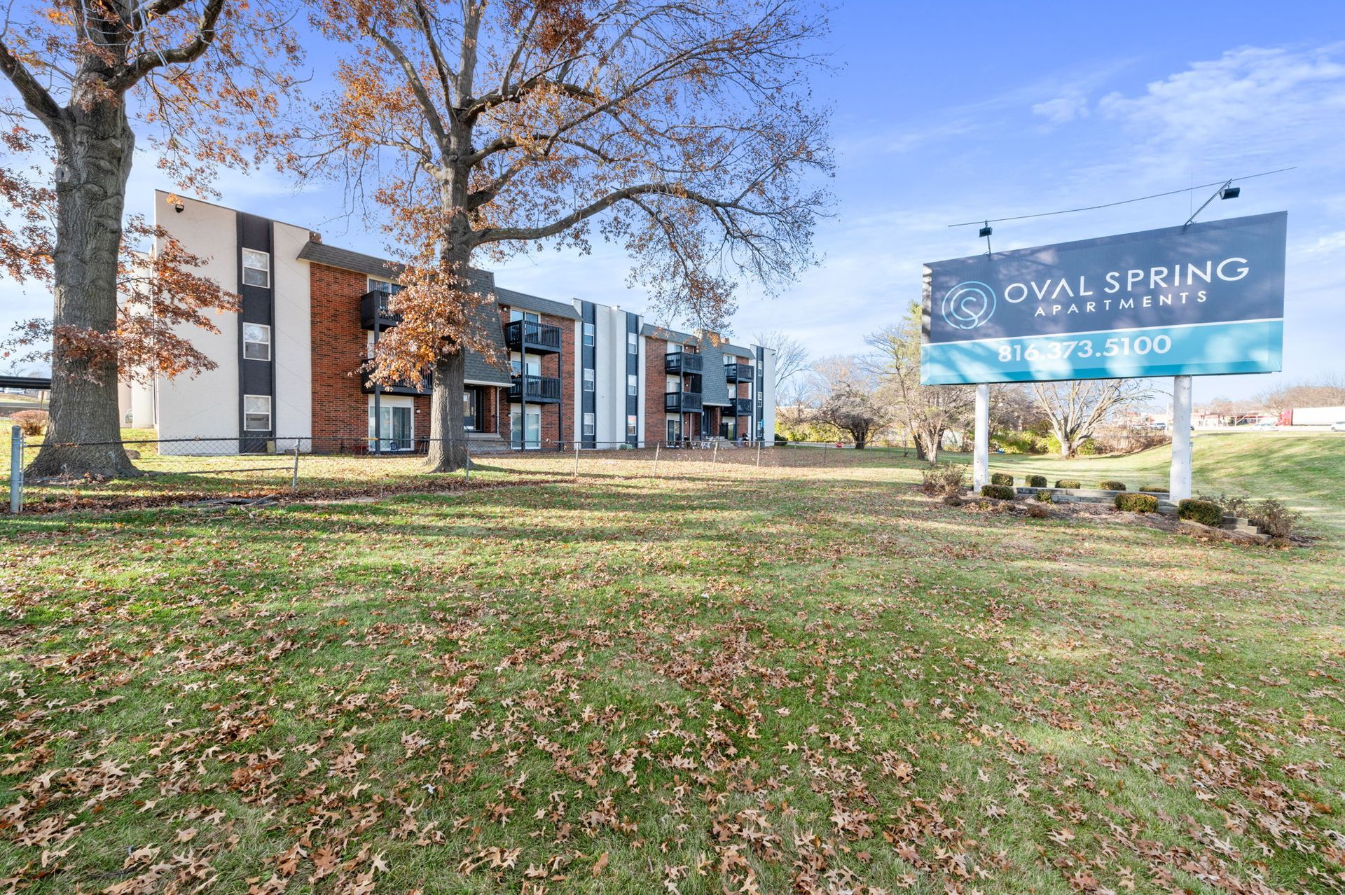 Apartment complex with blue sign and leaf-covered lawn under a tree in autumn