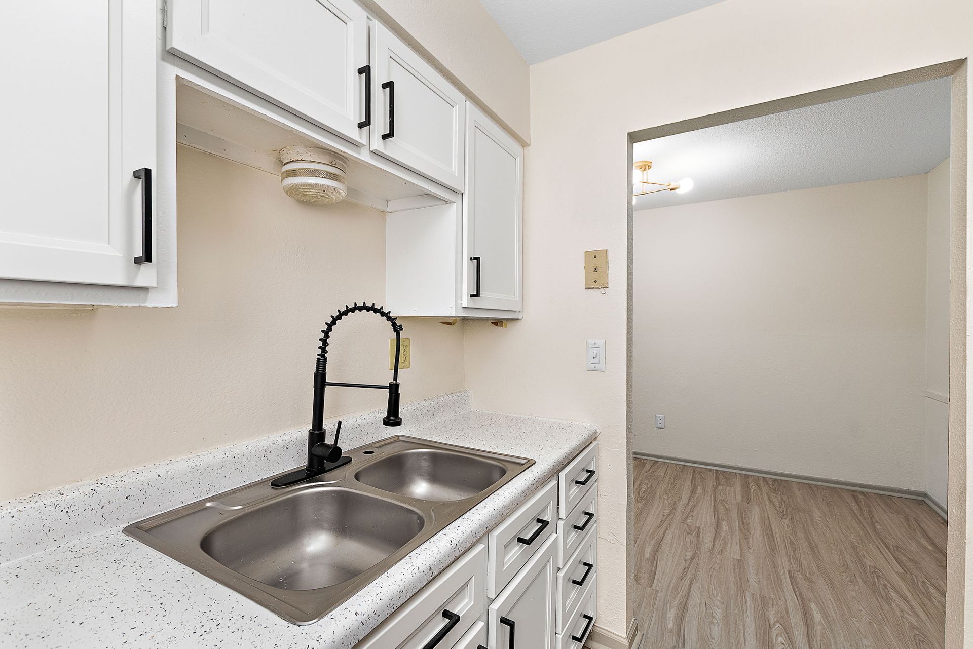 Small white kitchen with double sink, black faucet, and pass-through to an empty room with wood flooring