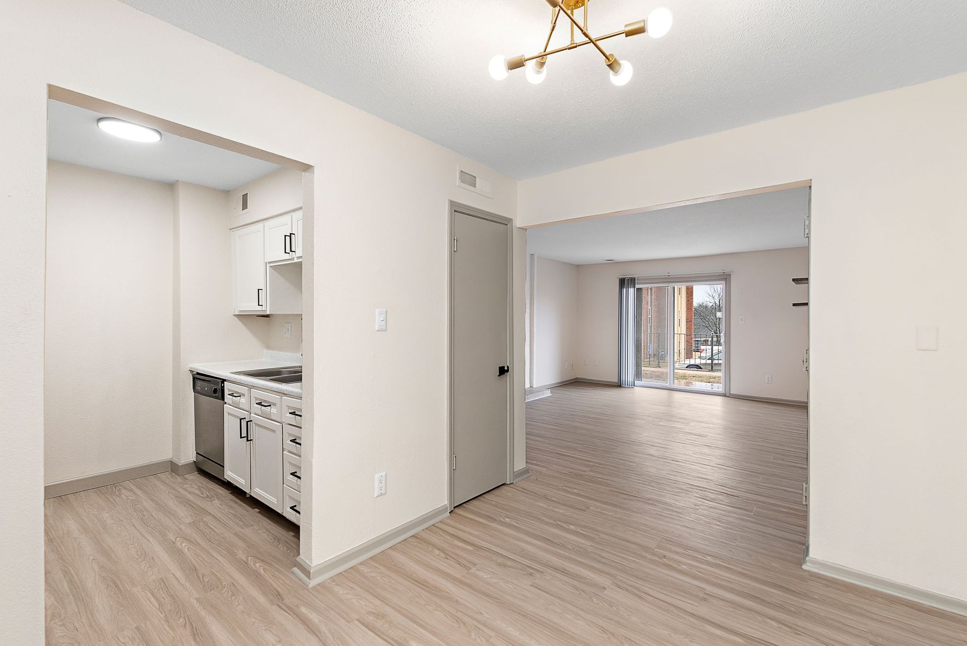 Empty apartment with light wood floors, white walls, kitchen nook, and open living area with chandelier.