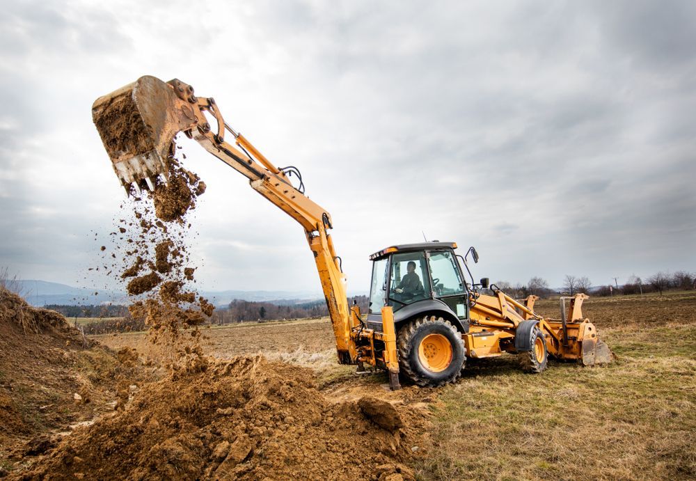 A man driving a yellow backhoe dumps soil on a field under an overcast sky.