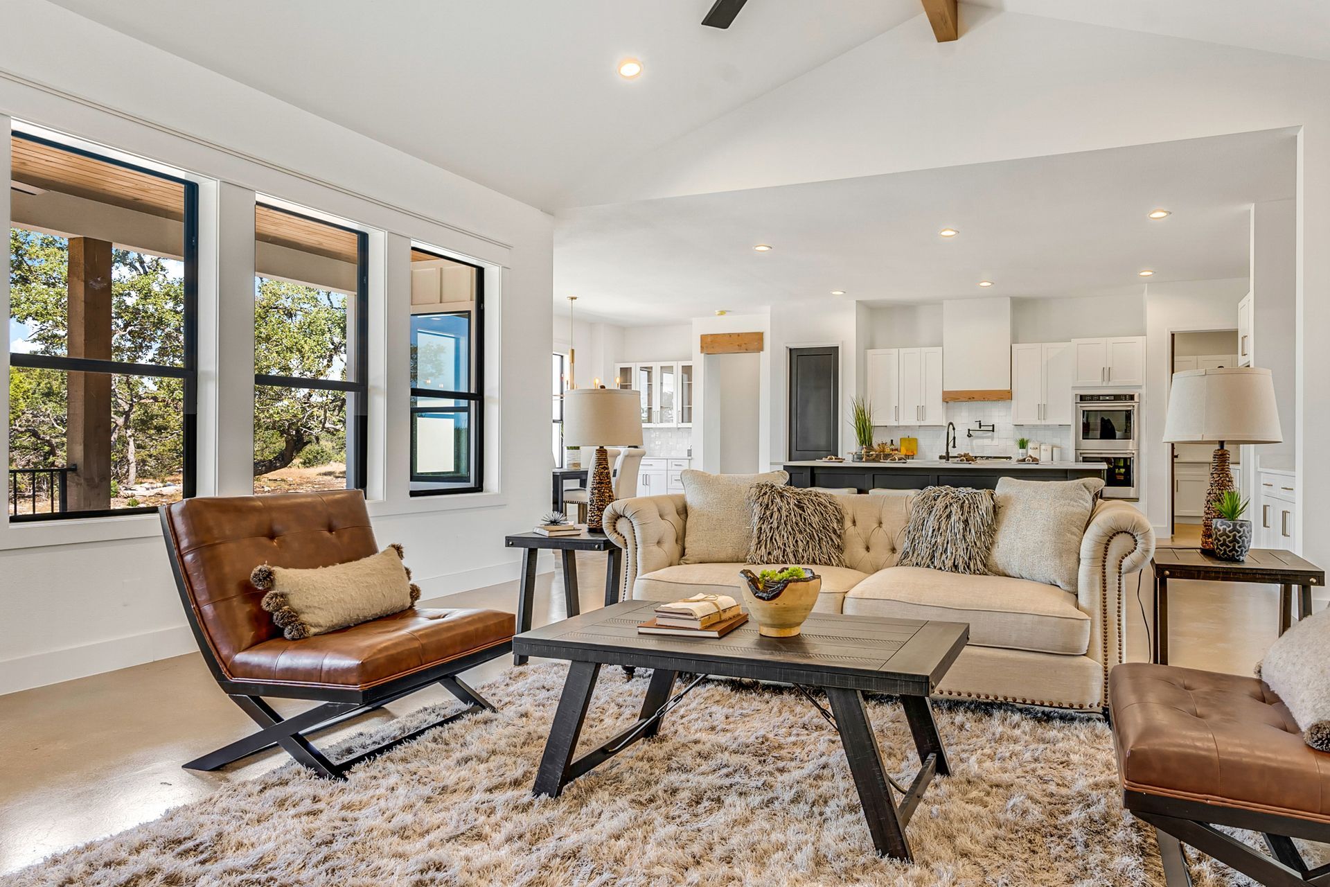 Bright modern living room with beige sofa, wood chairs, large windows, and open kitchen view