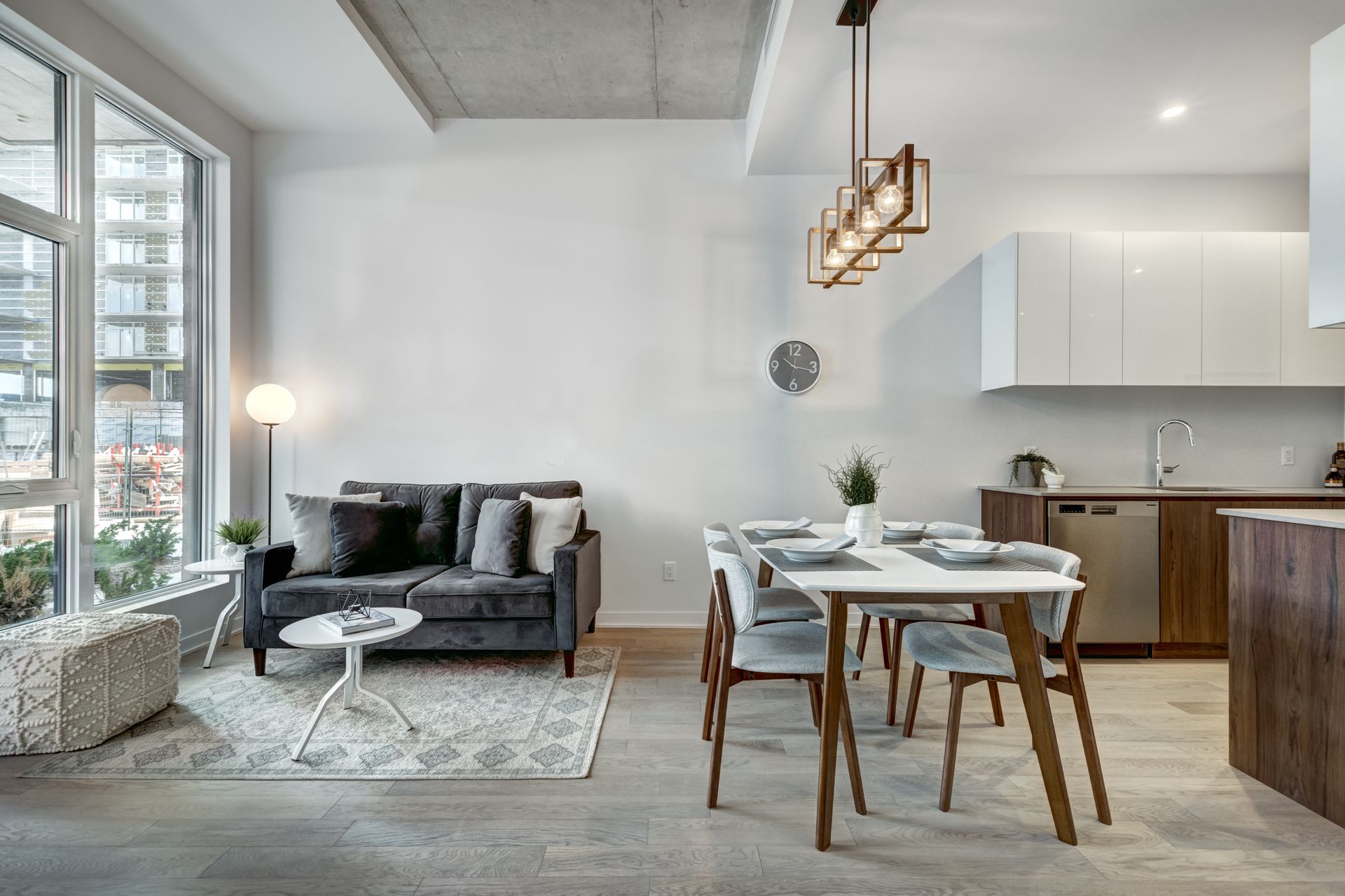 Modern open-plan living room and kitchen with gray sofa, dining table, pendant lights, and large windows