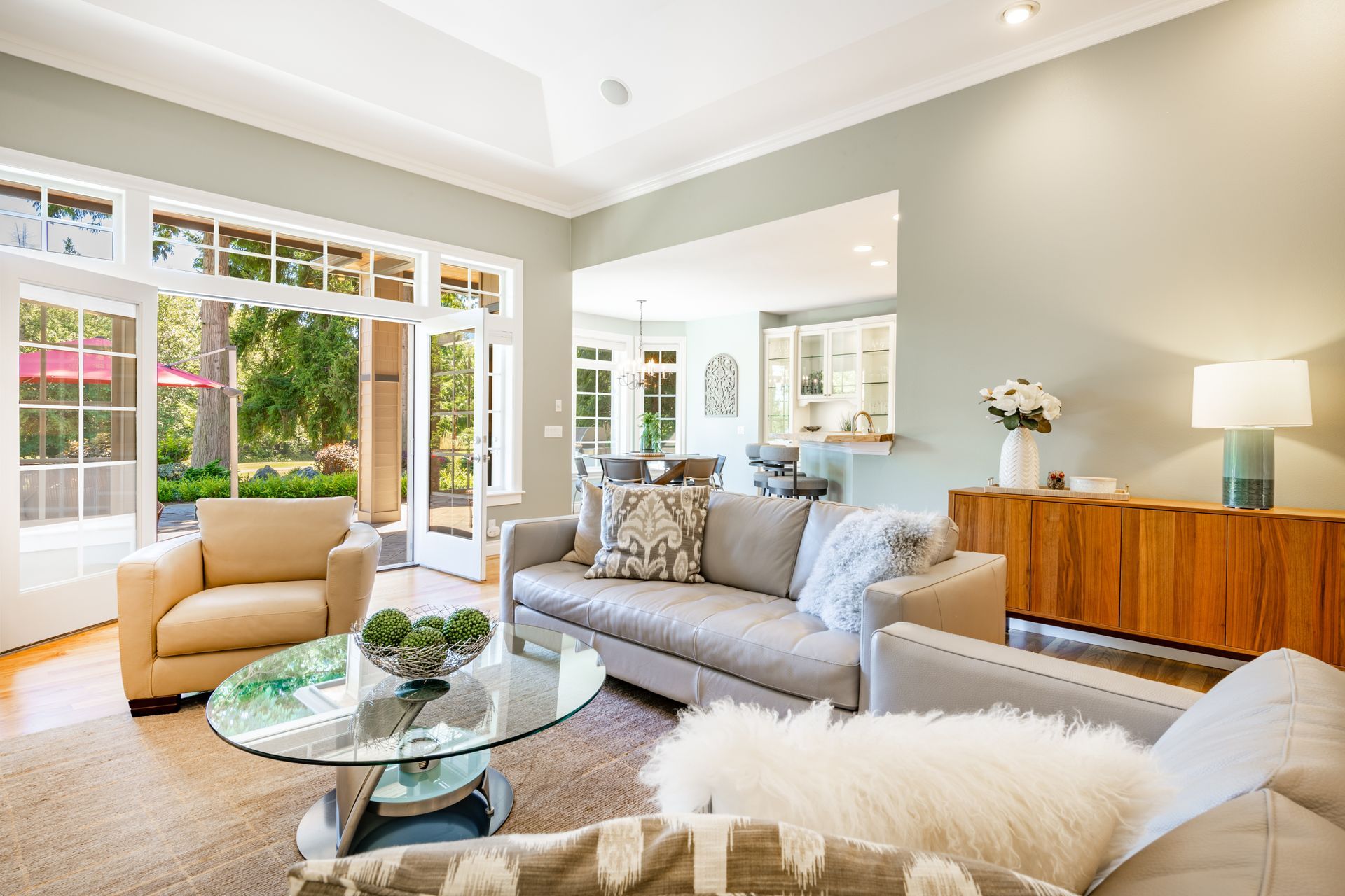 Bright living room with gray sofa, tan chair, glass coffee table, and large windows overlooking a garden