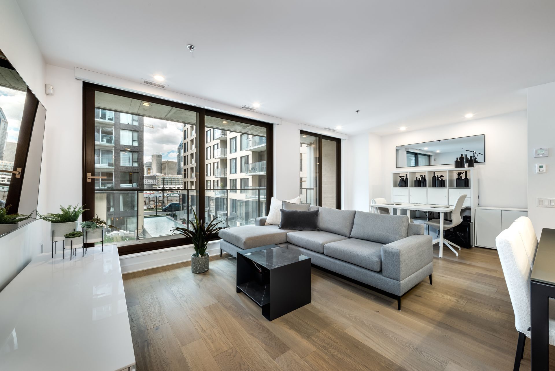 Bright modern living room with gray sofa, coffee table, large windows, and dining area.