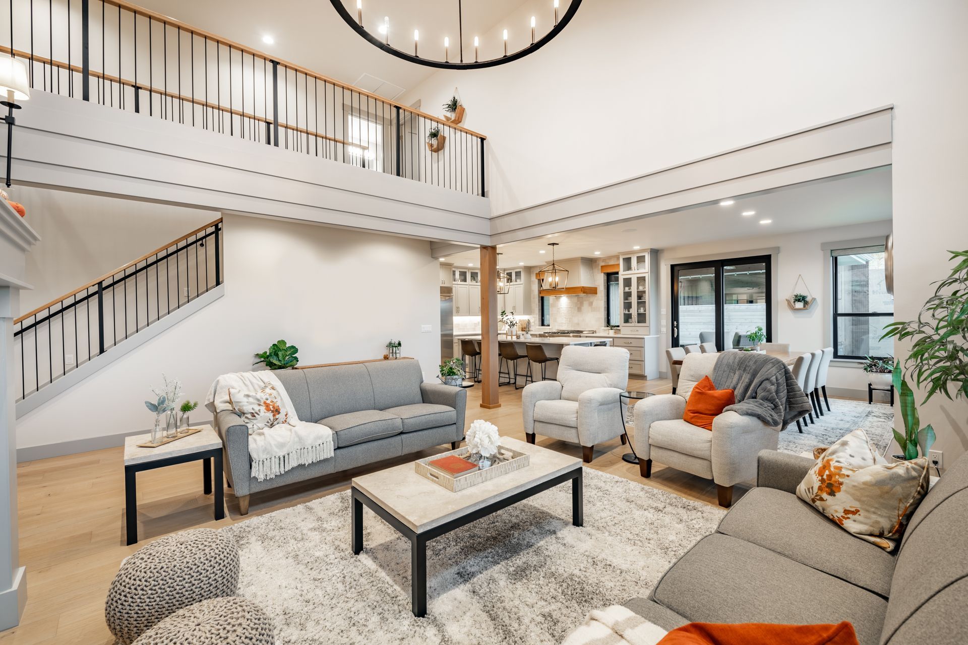 Modern living room with gray sofas, patterned rug, coffee table, staircase, and loft balcony under a circular chandelier