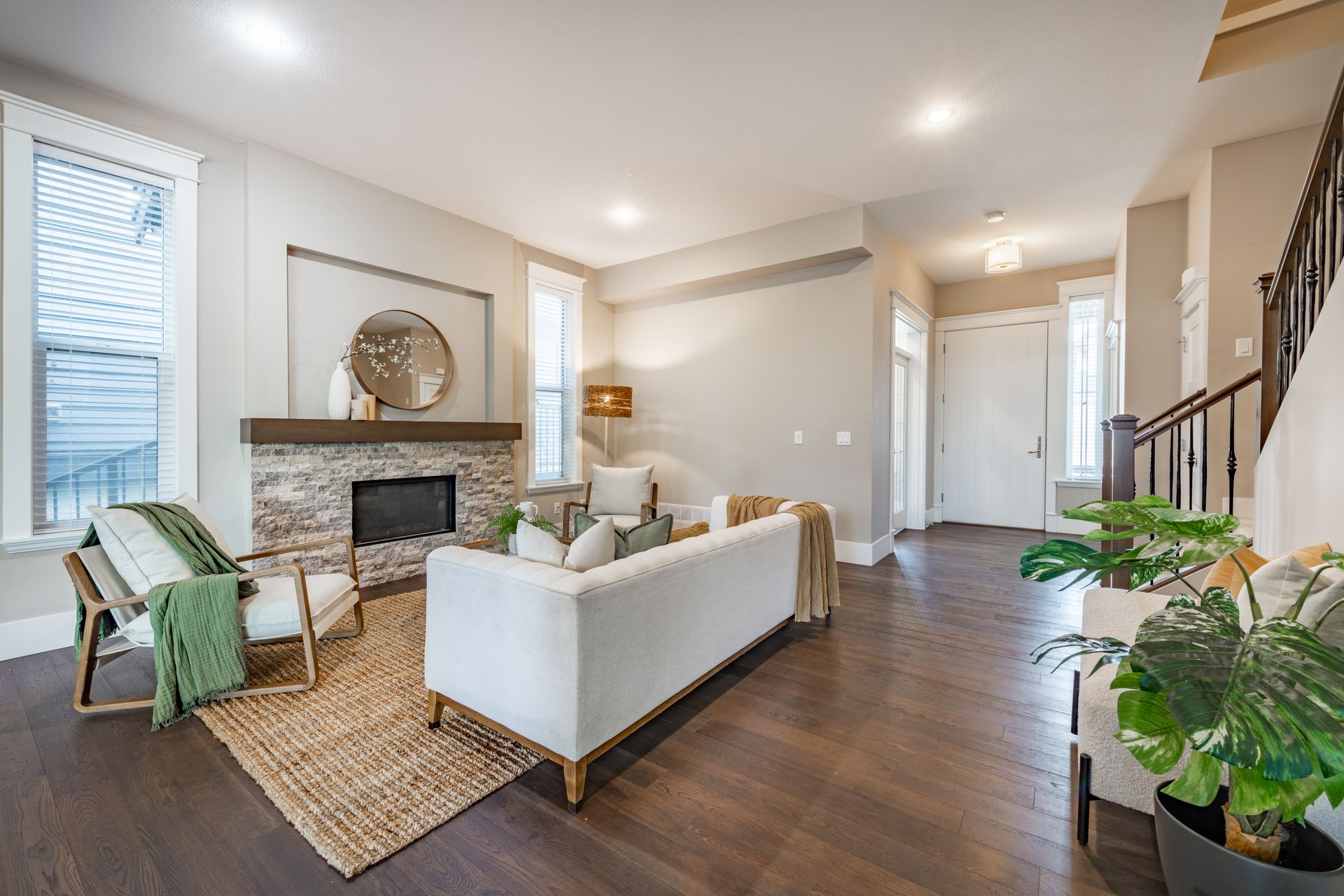 Bright living room with white sofas, fireplace, woven rug, dark floors, and staircase by hallway