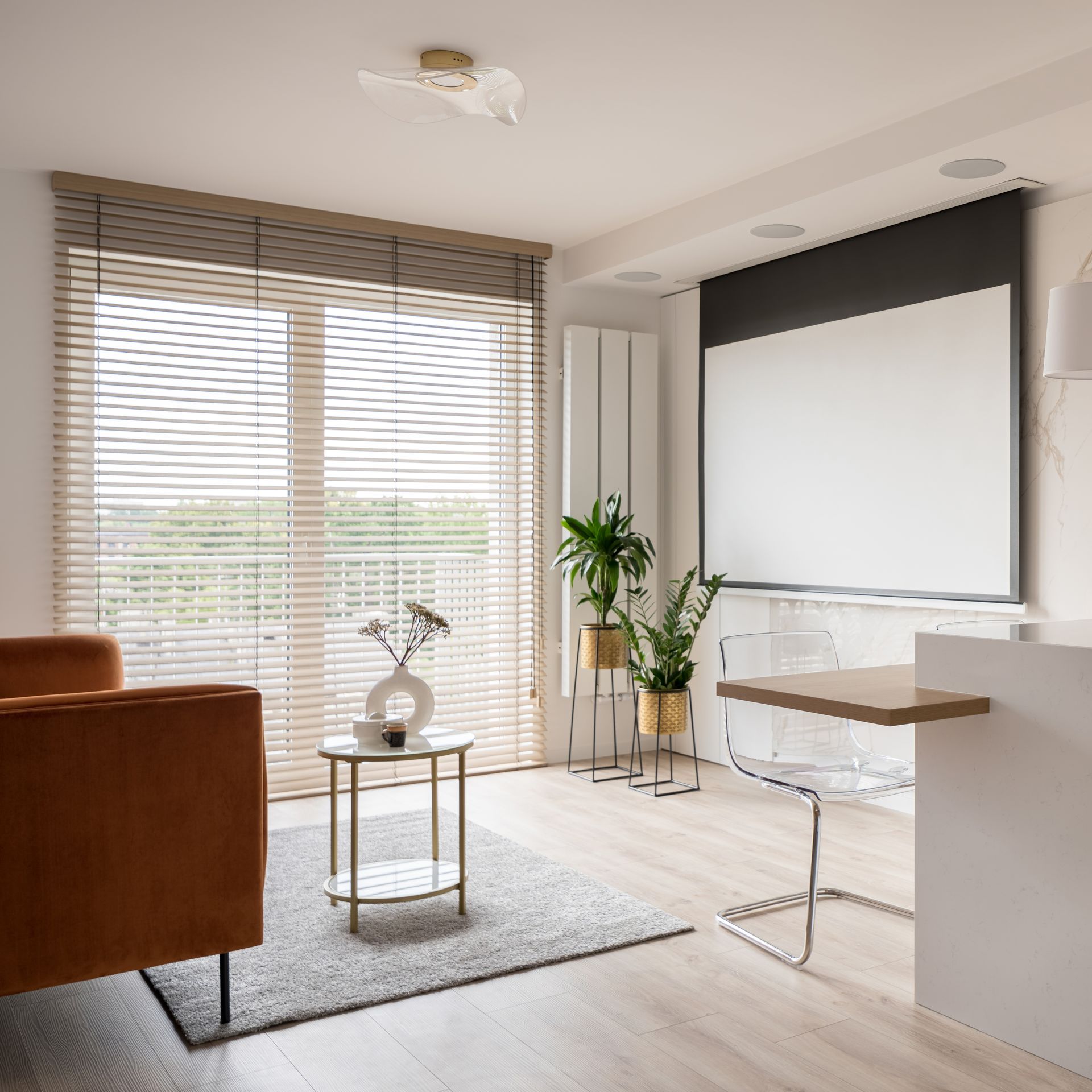 Bright modern living room with window blinds, projector screen, potted plants, and a small round table