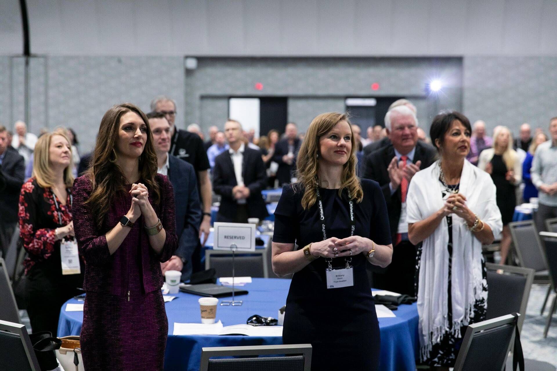 People at a conference clapping, some standing near a blue table, with many more attendees in the background.