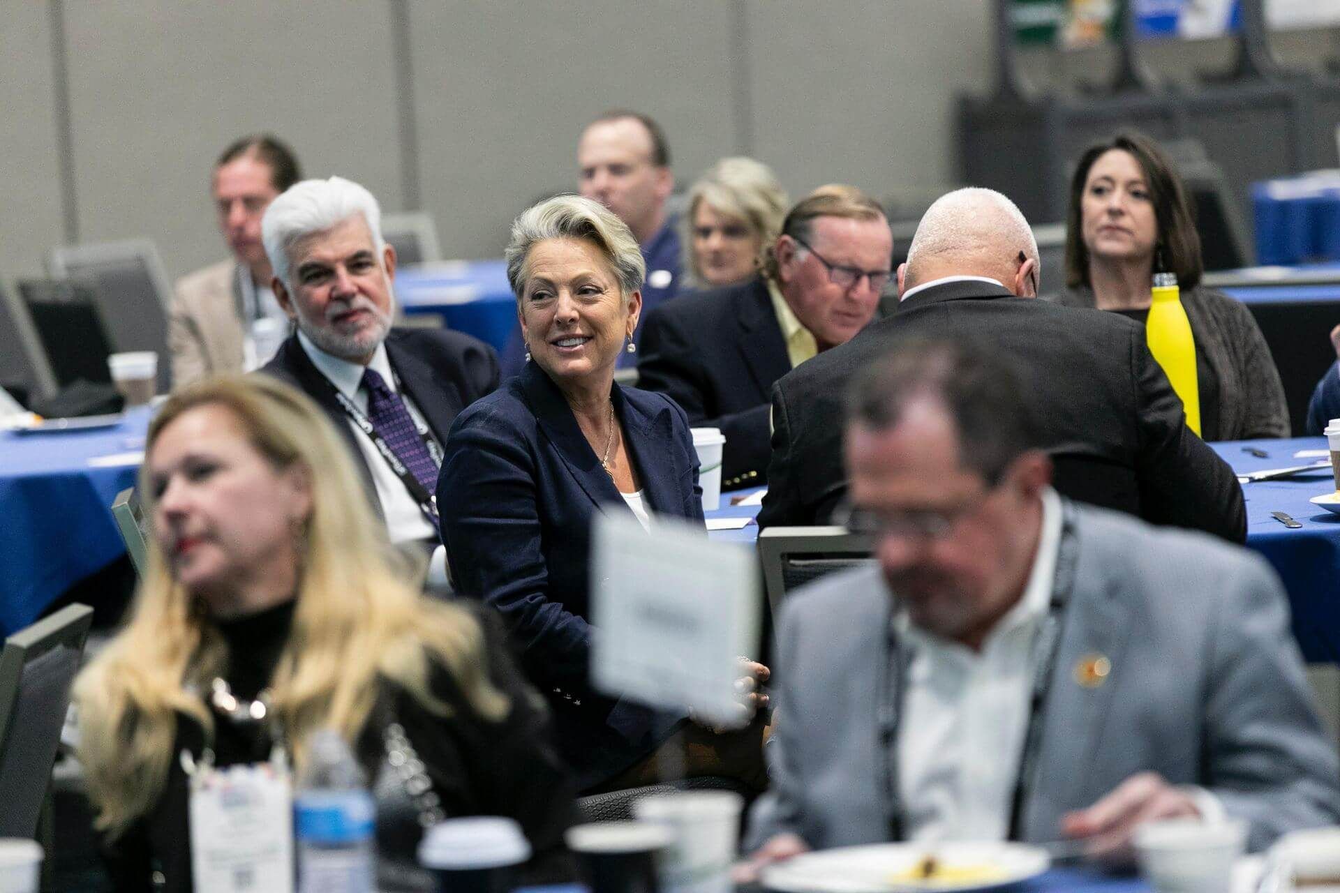 People at a conference seated around tables; a woman in a blue blazer smiles.