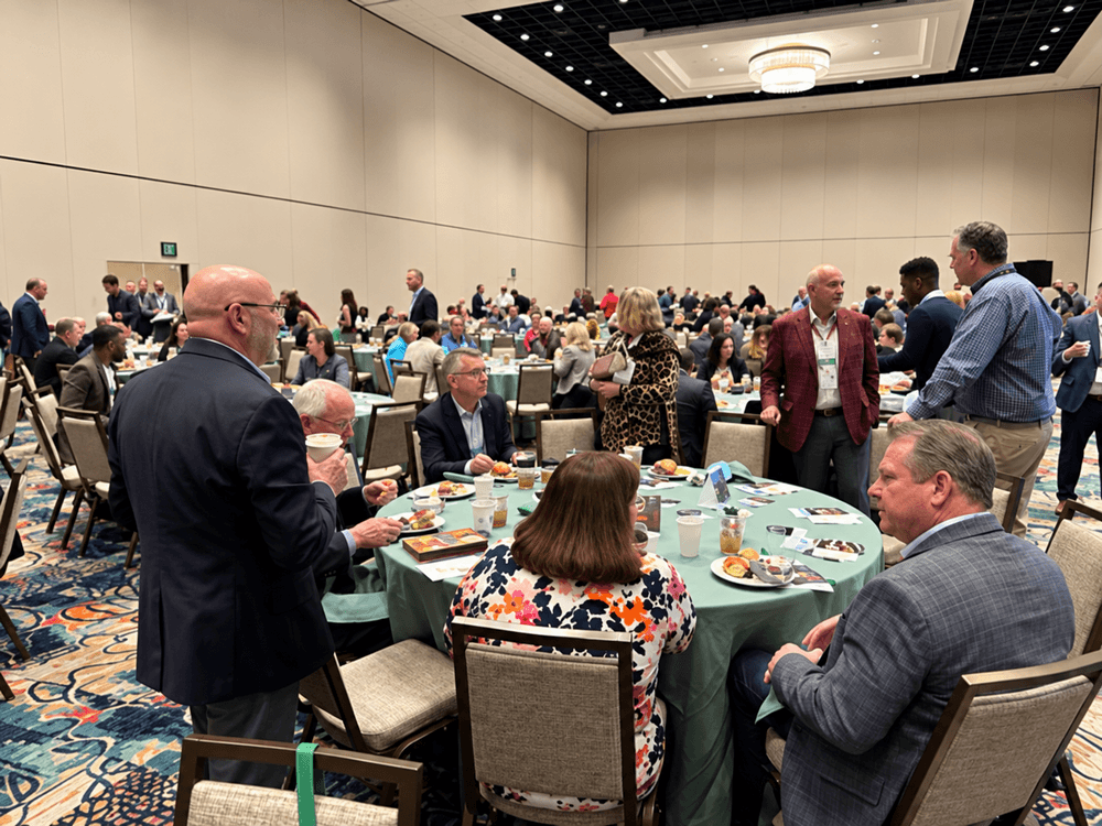 Large ballroom with tables, people seated, presentation screens, and a speaker at a podium.
