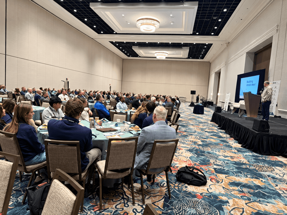Man speaking at a podium in a large conference hall with seated audience. Stage, monitors, tables, and carpet.