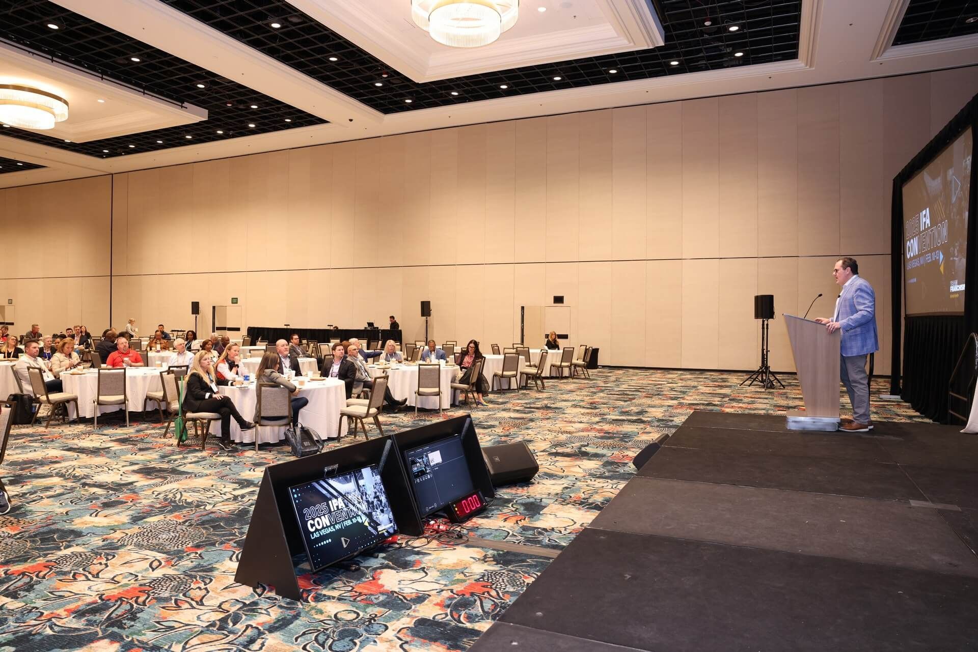 Man speaking at a podium in a large conference hall with seated audience. Stage, monitors, tables, and carpet.